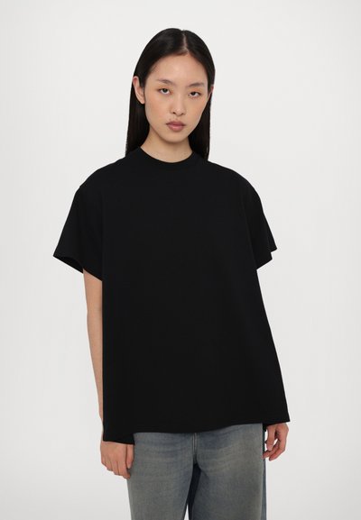 Juodas per didelis T-shirt su trumpomis rankovėmis, pagamintas iš minkštos medvilnės. Turi apvalų kaklą ir lygią tekstūrą. Dėvimas su šviesiai mėlynomis džinsais.
