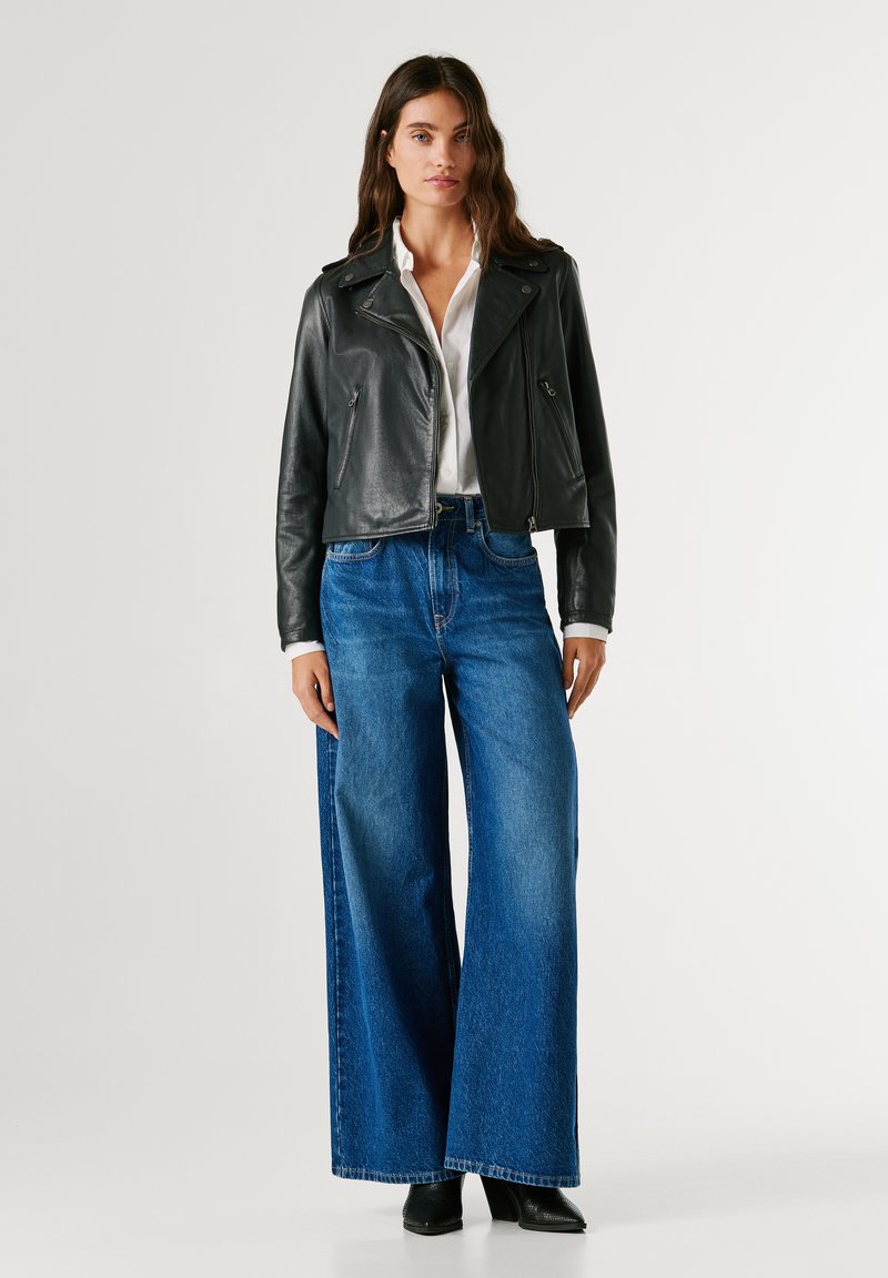 Μαύρο δερμάτινο κοντό μπουφάν με φερμουάρ, φορεμένο πάνω από ένα λευκό πουκάμισο. Παντελόνι με φαρδιά πόδια από μπλε τζιν και μαύρες μπότες ολοκληρώνουν το look.