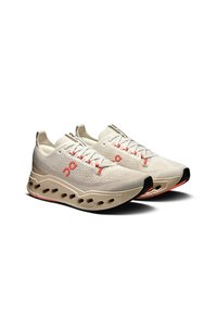 Light beige athletic shoes with a textured knit upper, red accents, and a sculpted sole featuring cut-out sections for cushioning.