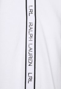 Vit tyg med en vertikal svart rand och präglad text som läser "RALPH LAUREN" och "LRL" i alternerande mönster.