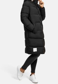 Svart puffjacka med huva, med en framdragkedja, sidofickor och en logotyp på fållen. Bärs över mörka jeans och vita sneakers.