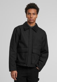 Young man with curly hair wearing a black jacket with large front pockets and a corduroy collar, standing against a plain gray background.