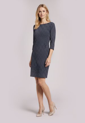 Femme debout portant une robe bleu marine au genou avec un motif géométrique et des chaussures à talons beiges sur un fond uni.