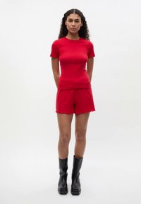 Vrouw met krullend haar, gekleed in een rood T-shirt met korte mouwen en bijpassende shorts, staand met de handen achter haar rug en zwarte laarzen tot halverwege de kuit.