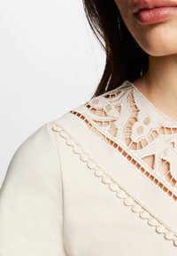 Top de crema claro con detalles de encaje en el escote, con patrones recortados y borde con ondas. Textura de tela suave.