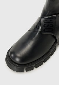 Stivale in pelle nera con suola chunky, caratterizzato da una larga fascia lucida con testo sulla parte superiore. Finitura liscia e design moderno.