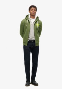 Felpa verde con cappuccio e lacci, con un logo anteriore, indossata sopra una maglietta grigia. Abbinata a jeans in denim scuro e scarpe nere. Design semplice.