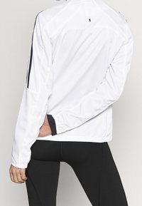 Chaqueta atlética blanca con detalles en negro, material ligero, mangas largas y una textura suave. Presenta un pequeño logo en la parte posterior.
