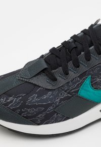 Chaussure de sport noire avec un tissu texturé, des accents gris et un logo Nike vert. Les lacets sont noirs ; la semelle est blanche avec un motif de crampons.