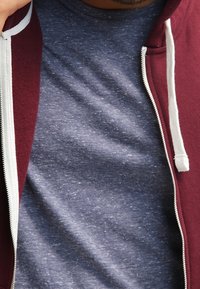 Burgundská mikina na zip s bílými tkaničkami, navrstvená přes texturované námořnické modré tričko, obě s uvolněným, pohodlným střihem.