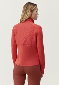 Punainen urheilu takki, jossa on pitkät hihat, korkea kaulus ja tikkurakenteinen kuusikulmainen muotoilu yläselässä; sileä kangas, joka on tiiviisti istuva.