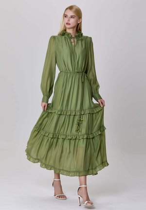 Femme blonde portant une robe maxi à manches longues de couleur olive avec des volants et un col fleuri, tenant l'ourlet, associée à des sandales à talons blancs.