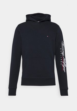 Hoodie noir avec poche avant, logo Tommy Hilfiger sur la poitrine gauche, et texte en script blanc avec accent rouge sur la manche droite.