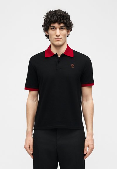 Άνδρας με σγουρά σκούρα μαλλιά, φορώντας μαύρο πόλο πουκάμισο με κόκκινο γιακά και άκρες μανικιών, στέκεται μπροστά σε απλό λευκό φόντο.