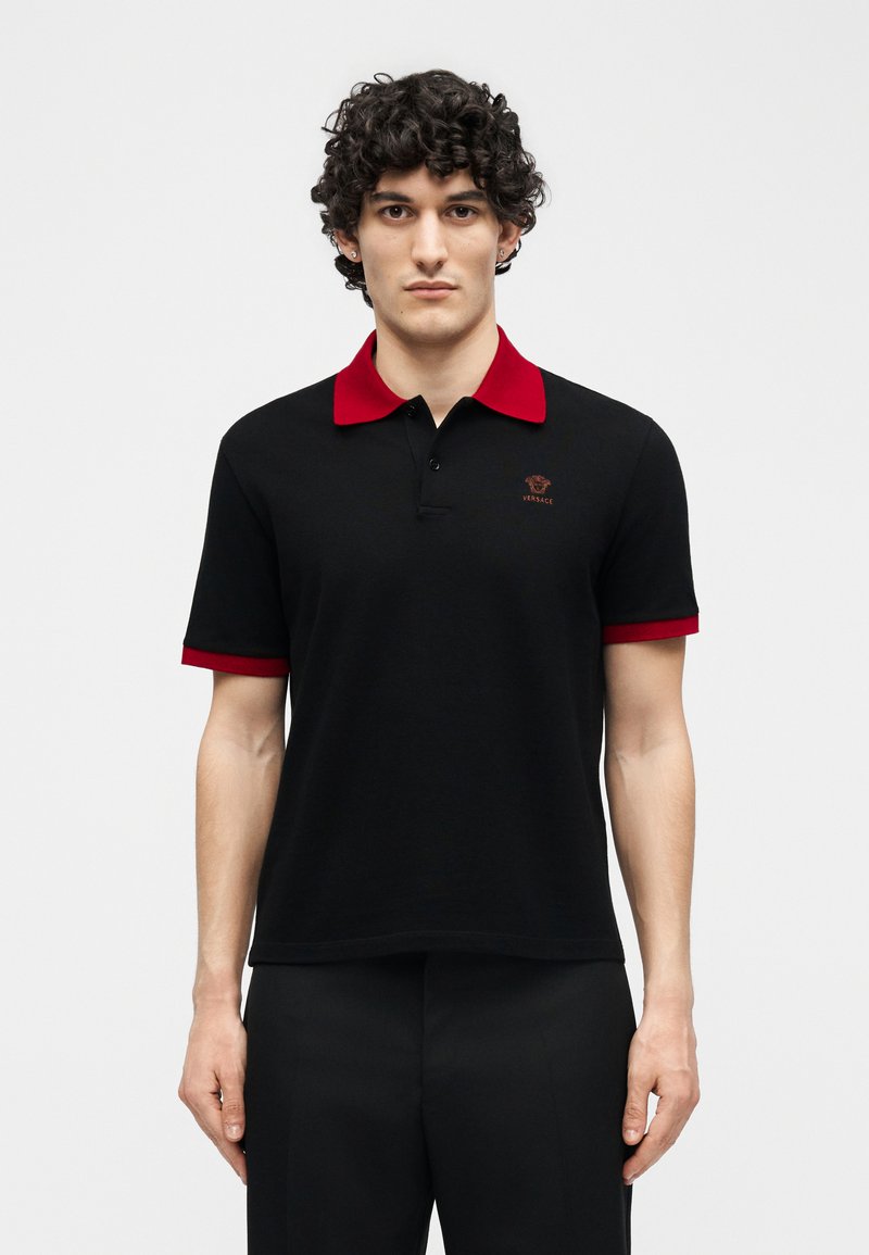 Man with curly dark hair wearing a black polo shirt with red collar and sleeve edges, standing against a plain white background.