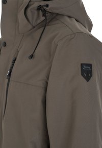 Brun vattentät jacka med huva, med en kontrasterande svart dragkedjeficka och en badge på ärmen. Texturerad tyg och justerbar dragsko.