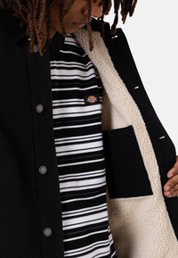 Veste en velours côtelé noir avec un intérieur en laine, accompagnée d'une chemise rayée noir et blanc, de boutons argentés et d'une petite étiquette de logo.
