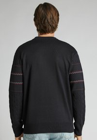 Ciemnogranatowy sweter z dzianiny, z prążkowanym okrągłym dekoltem, rękawami w warkoczowy splot oraz kontrastującymi, czerwono-białymi zygzakowatymi paskami na górnych partiach ramion.