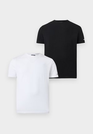 Άνδρας που στέκεται ξυπόλητος φορώντας εφαρμοστό μαύρο κοντομάνικο πουκάμισο και μαύρα σλιπ μπόξερ σε απλό φωτεινό φόντο.