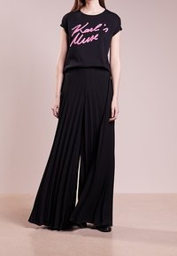 Černé tričko s růžovým nápisem "Karl's Muse" v kombinaci s černými širokými kalhotami s plisé. Hladká látka a uvolněný střih.