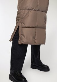 Gilet imbottito marrone con sezioni trapuntate, chiusure laterali a scatto, abbinato a jeans neri a gamba larga e stivali neri massicci.