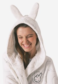 Hvid, blød hættebadekåbe med kaninører. Har en blød tekstur, broderede detaljer og et rundt grafisk motiv på brystet.