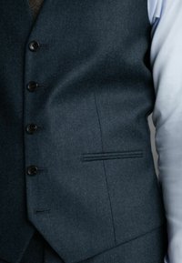 Dunkelgraue Wollweste mit fünfknöpfigem Verschluss, strukturierter Webart und einer seitlichen Tasche. Kombiniert mit einem hellen Hemd.