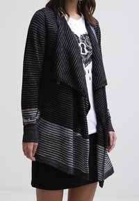 Svart- och grårandig cardigan med öppen framsida, draperad design och långa ärmar, tillsammans med en grafisk vit t-shirt.