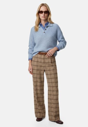Donna che indossa pantaloni marroni a quadri a gamba larga, maglione di lana celeste sopra camicia blu con colletto, cintura marrone, occhiali da sole e scarpe scure, in piedi.
