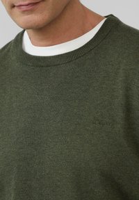 Dunkelgrüner Strickpullover mit Rippenkragen, darunter ein weißes T-Shirt mit Rundhalsausschnitt und einem dezenten Logodetail.