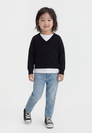 Παιδί με μαύρα μαλλιά μέχρι τους ώμους στέκεται φορώντας μαύρο πουλόβερ με V-λαιμόκοψη, λευκό πουκάμισο από κάτω, ανοιχτό μπλε τζιν και μαύρα αθλητικά παπούτσια.