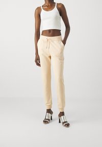 Pfirsichfarbene Trainingshose mit Kordelzugbund und Seitentaschen, kombiniert mit einem weißen, kurzen Tanktop und silbernen Absatzsandalen.