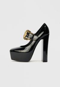 Zapatos de tacón alto de charol negro con plataforma y tacón grueso. Cuenta con una hebilla decorativa dorada y un diseño de puntera redondeada.