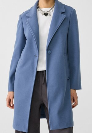 Blauwe wollen jas met één knoop, gedragen over een wit shirt en donkere broek, gecombineerd met een zilveren ketting met hartvormige hangers.