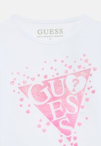Biały bawełniany T-shirt z różowym brokatowym wzorem w trójkątnym kształcie z napisem "GUESS" oraz sercami o różnych rozmiarach.