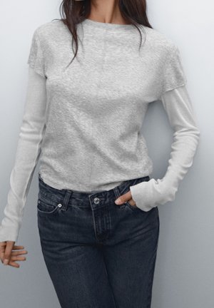 Femme portant des hauts gris superposés à manches longues et courtes avec un jean taille haute foncé, une main dans la poche, sur un fond uni.