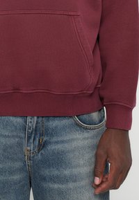 Felpa bordeaux con tasca frontale e polsini a costine, abbinata a jeans blu sbiaditi. Il materiale appare morbido e testurizzato.