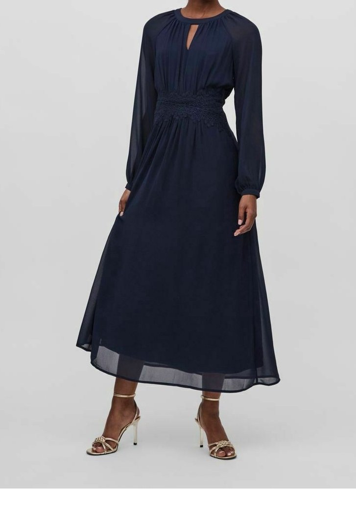 Femme portant une robe midi bleu marine à manches longues avec taille smockée et encolure en trou de serrure, associée à des sandales dorées à talons hauts.