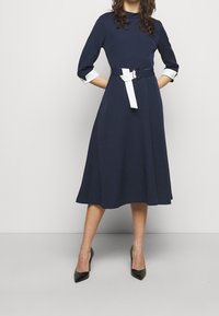 Marineblaues Midikleid mit Dreiviertelärmeln, ausgestattet mit einem kontrastierenden weißen Sash-Gürtel und einem ausgestellten Rock. Glatte Textur, lässiges Design.