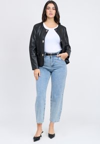 Giacca di pelle nera con bottoni a pressione, abbinata a una t-shirt bianca e jeans a vita alta di un blu chiaro. Scarpe nere a punta. Sfondo semplice.