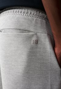 Hellgraue Jogginghose mit strukturiertem Muster, ausgestattet mit einem elastischen Bund, einer Seitentasche und einem kleinen geprägten Logo auf der Tasche.