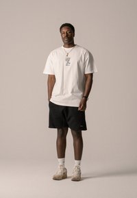 White cotton t-shirt with graphic print and rounded neckline, paired with black shorts, white ankle socks, and beige sneakers.
