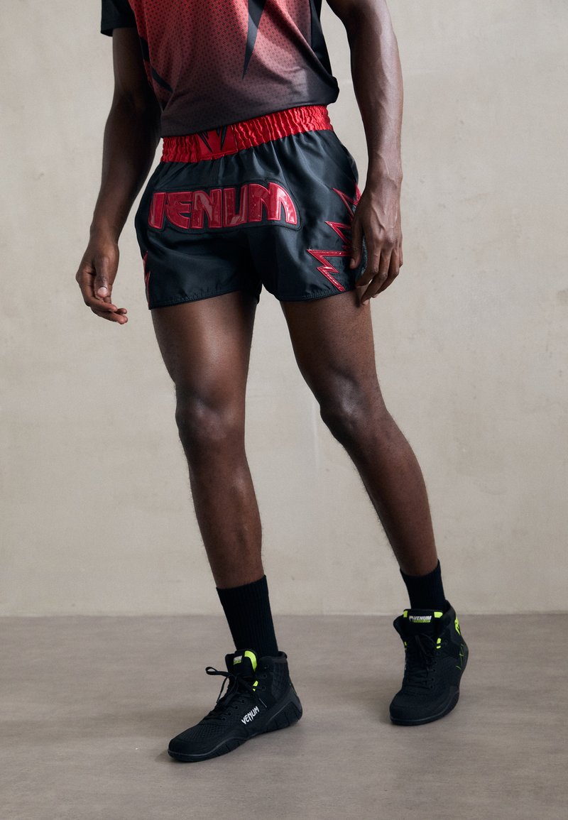 Schwarze Sportshorts mit rotem Schriftzug und Blitzmuster, kombiniert mit schwarzen High-Top-Sneakern mit Neonakzenten.
