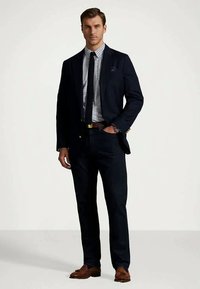 Mężczyzna ubrany w ciemnoniebieski garnitur, w kratkę koszulę, ciemny krawat, brązowy pasek i brązowe mokasyny, stoi z jedną ręką w kieszeni na jednolitym tle.