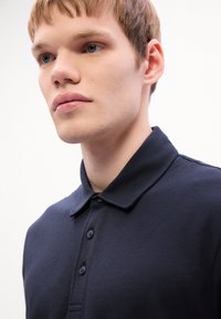 Joven con cabello corto castaño claro, vistiendo una camiseta polo de color azul marino oscuro, mirando ligeramente hacia la izquierda sobre un fondo blanco liso.