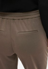 Brązowe, rozciągliwe spodnie z tkaniny o gładkiej fakturze, wyposażone w marszczony, elastyczny pas w talii oraz dwie tylne kieszenie.