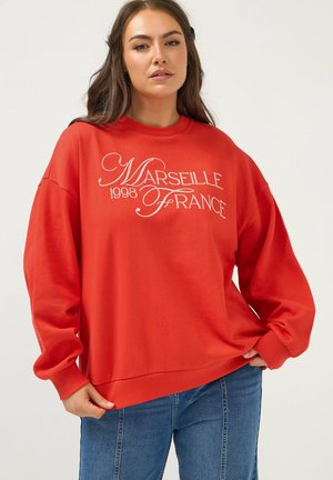 Frau trägt roten Pullover mit dem weißen Schriftzug "Marseille 1998 Frankreich", kombiniert mit blauer Jeans, steht vor einem schlichten Hintergrund.