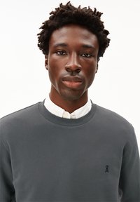 Grijze sweatshirt met een gestructureerde afwerking, ronde halslijn en klein logo op de linkerborst, gedragen over een witte blouse met kraag.