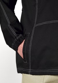 Personne portant une veste noire avec des coutures blanches, main reposant dans la poche latérale, pantalon beige visible en bas.