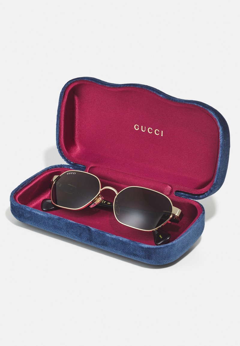 Gucci sunglasses inside a navy velvet case with a burgundy interior. Sunglasses feature dark lenses, gold frames, and black tips.